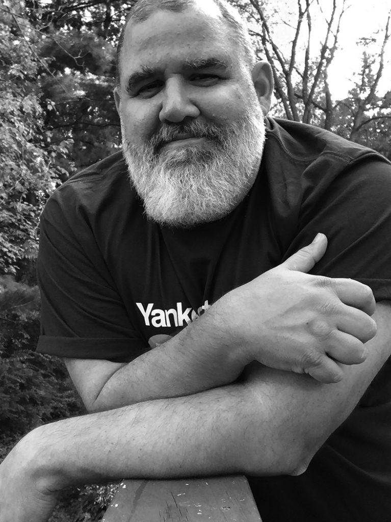 Portrait of Kenneth Carozza, a man with a short-cropped haircut and full white beard, smiling softly while leaning forward with arms crossed on a surface, wearing a dark t-shirt, outdoors with trees in the background, in black and white