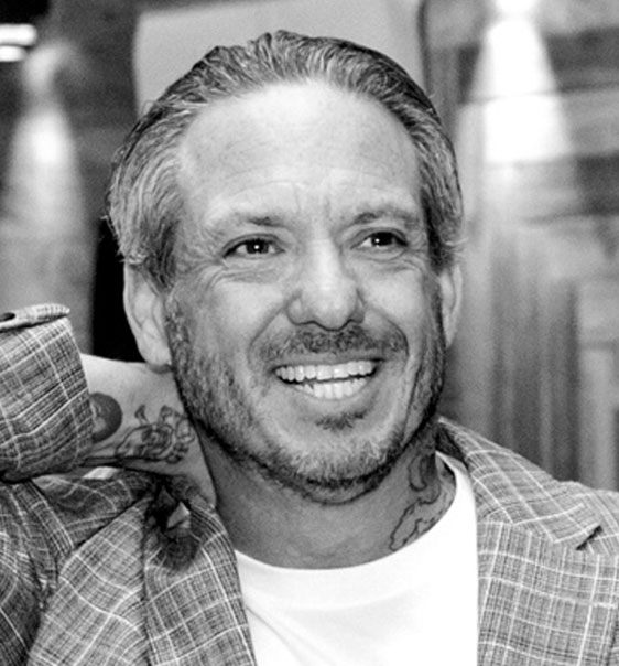Black-and-white headshot of Brian Zandman smiling, gray hair, neck tattoo, checkered blazer
